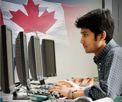 Indian student working in front of computers with Canadian flag in the background.