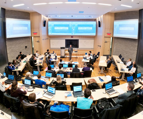 Students sit in a large lecture hall while listening to the professor at the front of the room.