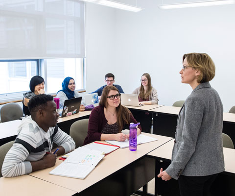A female professor addresses a classroom of diverse university students.