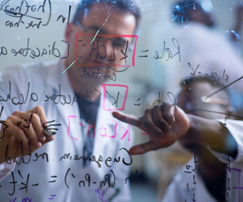 Two researchers writing math formulas on a plexiglass board.