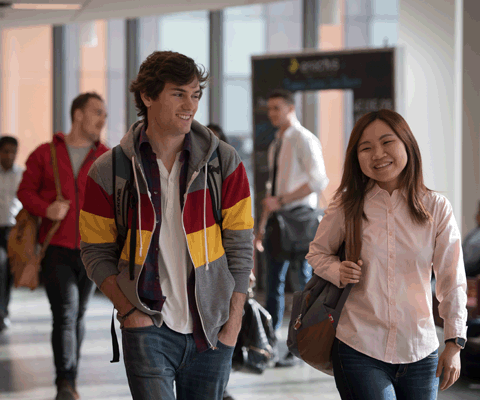 Two students walk down a university hallway together smiling and holding their Bags 