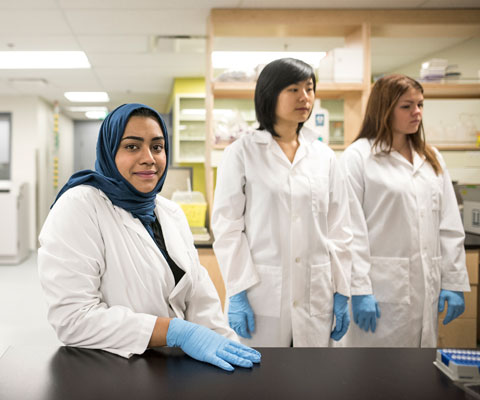 Trois chercheuses portant des blouses de laboratoires blanches sont debouts dans un laboratoire.