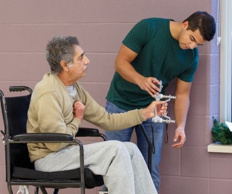 Male student testing a hand strength instrument with an elderly man in a wheelchair.