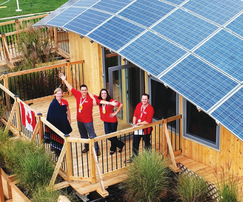 Four people standing on the balcony of a building with a rooftop covered entirely in solar panels.