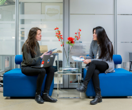 Two female students sit talking to one another in an office setting.