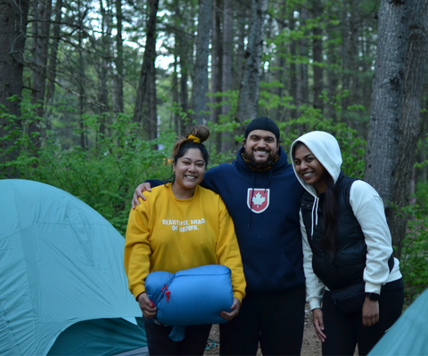 Three smiling students camping in the forest