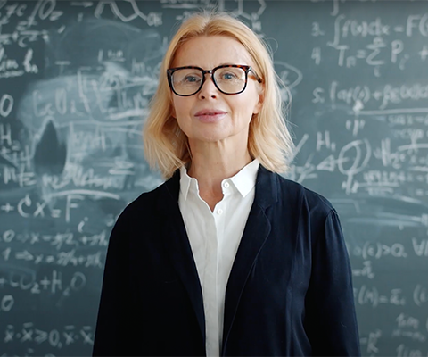 Middle-aged woman with glasses and professional clothing stands in front of a chalk board filled with equations