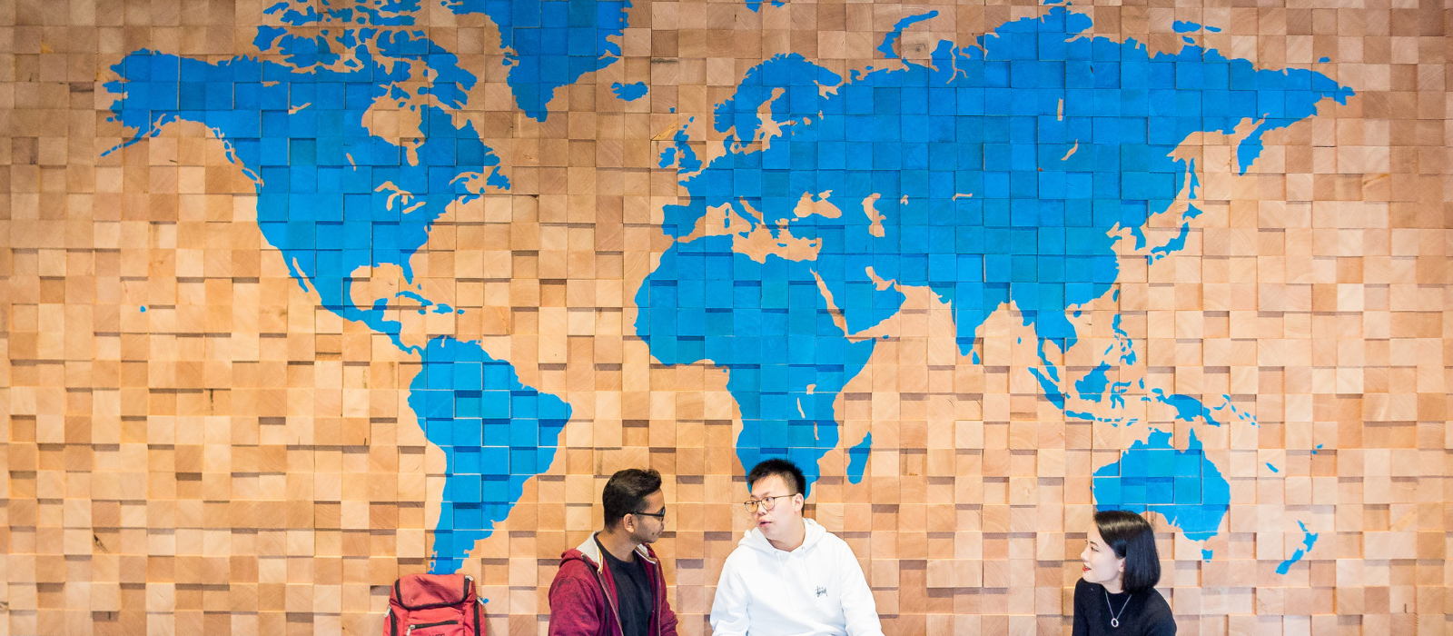 Three students sitting in front of a wall with a world map / 