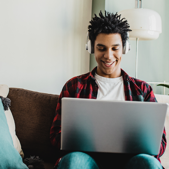 Student on couch surrounded by pillows wearing headphones and working on laptop