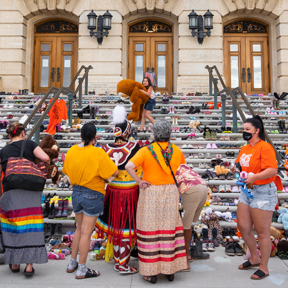 People wearing orange and traditional Indigenous clothing looking at the building's steps filled with children's shoes