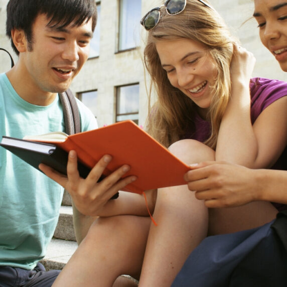 Three students sit on the steps outside a university building looking at the screen of an iPad and smiling.