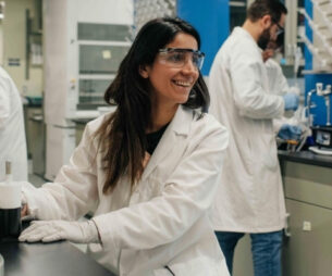 A student researcher wearing a white lab coat and safety goggles sits smiling in a university laboratory.