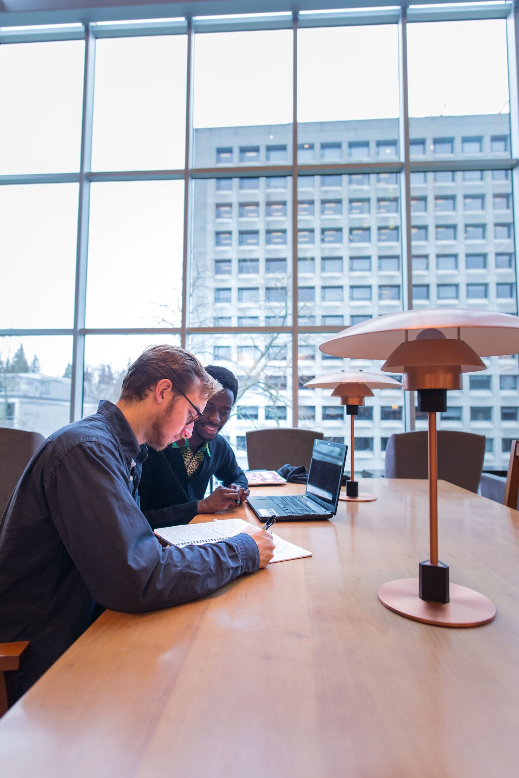 Two students studying at a table with a laptop and pen and paper