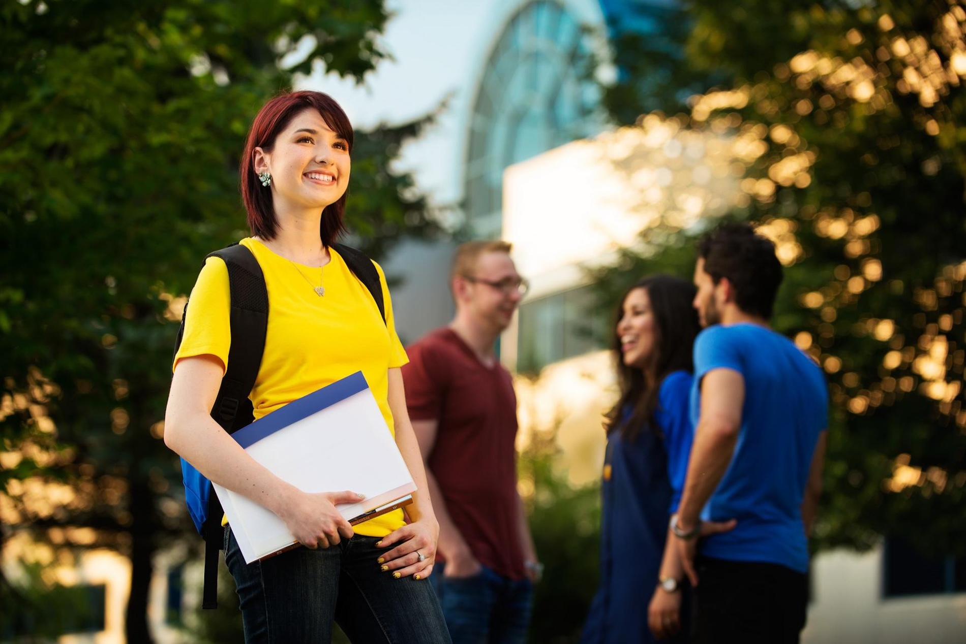 A student wearing a yellow shirt, wearing a backpack and holding notebooks smiling and posing with three students talking and laughing behind her