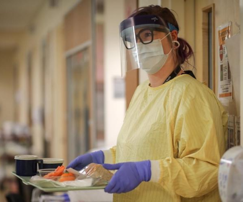 A person wearing protective gown and face shield holding a tray of food.