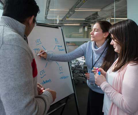 student writing on a white board with two other students looking on.