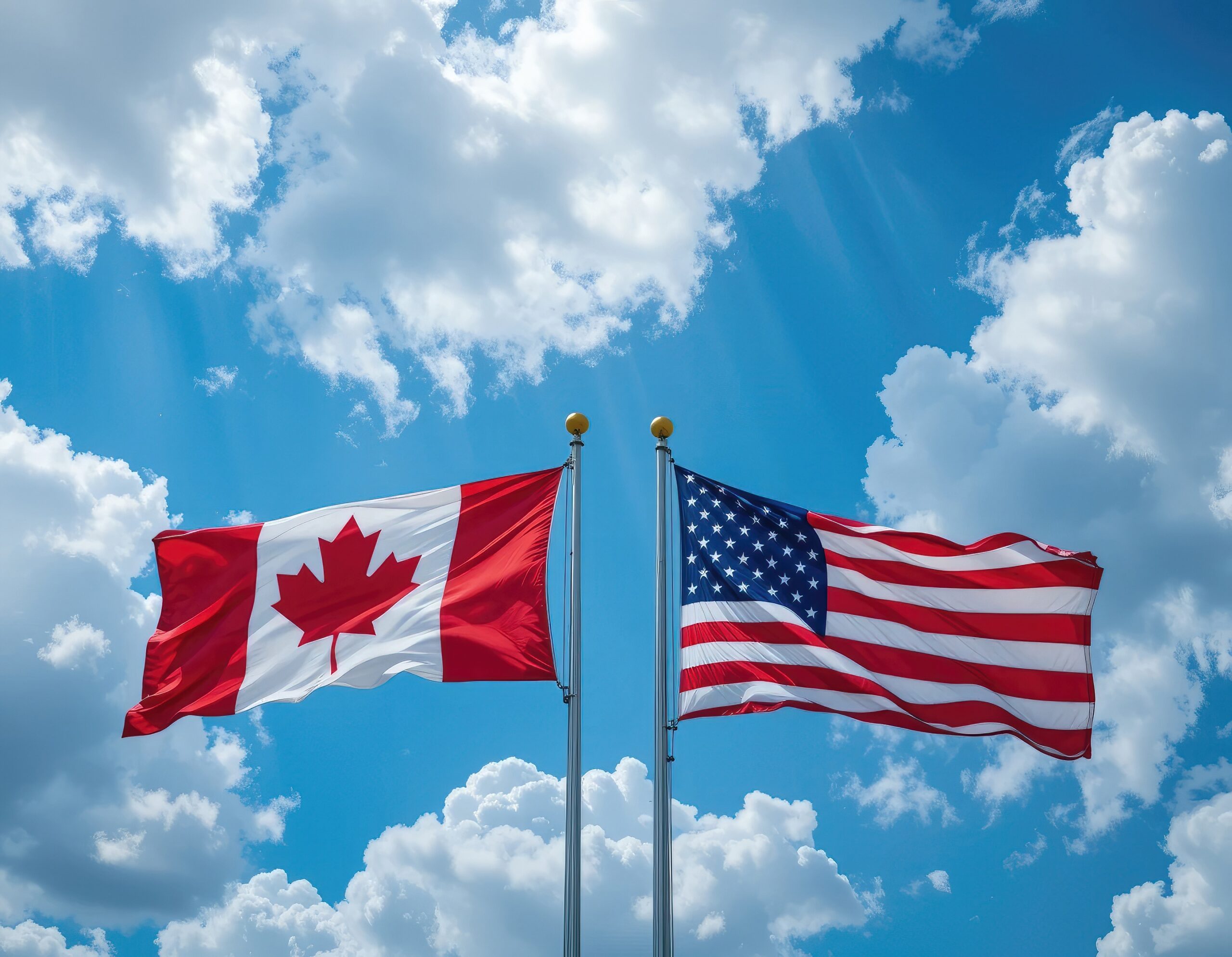 Des drapeaux américains et canadiens flottent dans le vent sur un ciel bleu vif avec des nuages ​​duveteux.