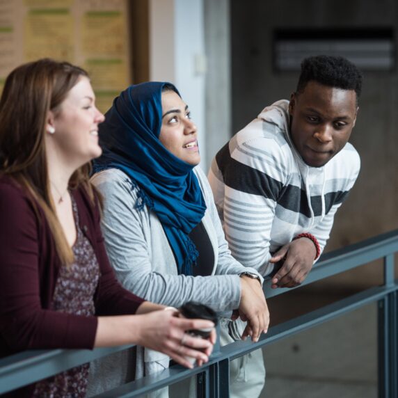 A group of three diverse students leaning over a railing and talking.